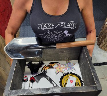 Axe Play staff member holding the Fun Box filled with throwing items including shovel, ninja stars, playing cards, spoons, and specialty axes